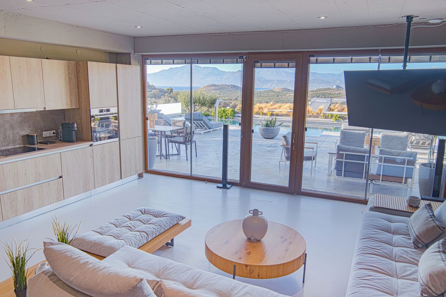 A spacious living area with a kitchen, a round wooden coffee table, and a large sliding glass door leading to an outdoor patio with seating and a pool.