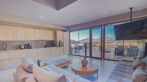 A modern living room with a large sofa, a coffee table, and a kitchen area with wooden cabinets and a flat-screen TV. - Marnos Grand Villa (Faistos)