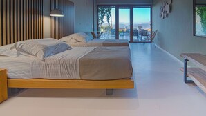 A modern bedroom with a large bed, a wooden headboard, and a view of the outdoors through large sliding doors. - Marnos Grand Villa (Faistos)