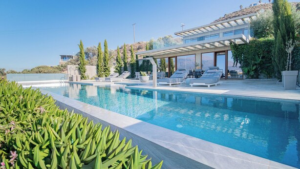A modern pool area with lounge chairs, a covered seating area, and a view of the surrounding landscape. - Marnos Grand Villa (Faistos)