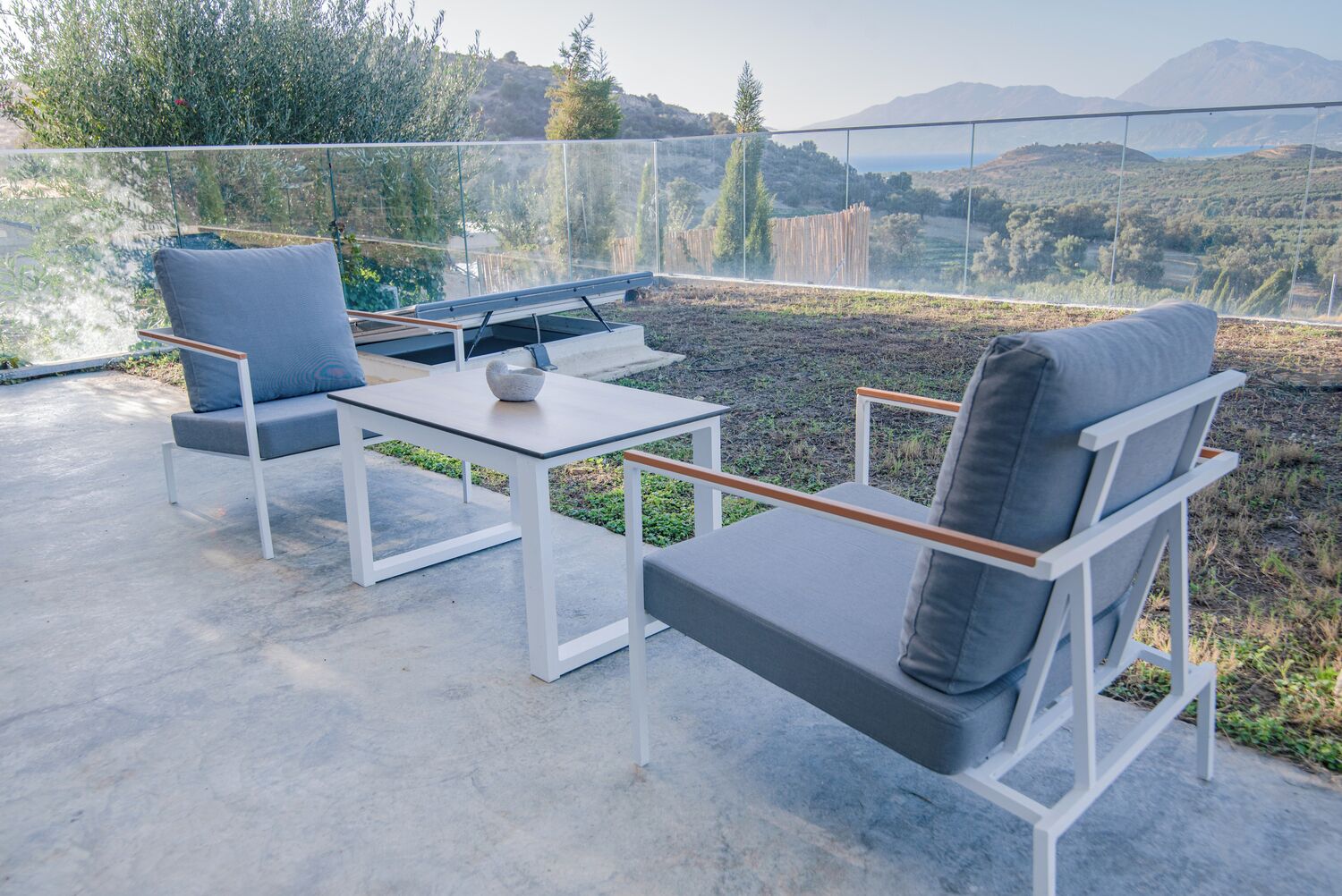 Un espace extérieur aménagé avec une table et deux chaises, offrant une vue sur un paysage pittoresque composé de collines et d’arbres.