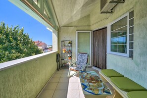Ein Balkon mit grüner gepolsterter Bank, einem kleinen Tisch mit bunten Untersetzer, einem Metallregal mit Topfpflanzen und einem Fenster mit Jalousien.