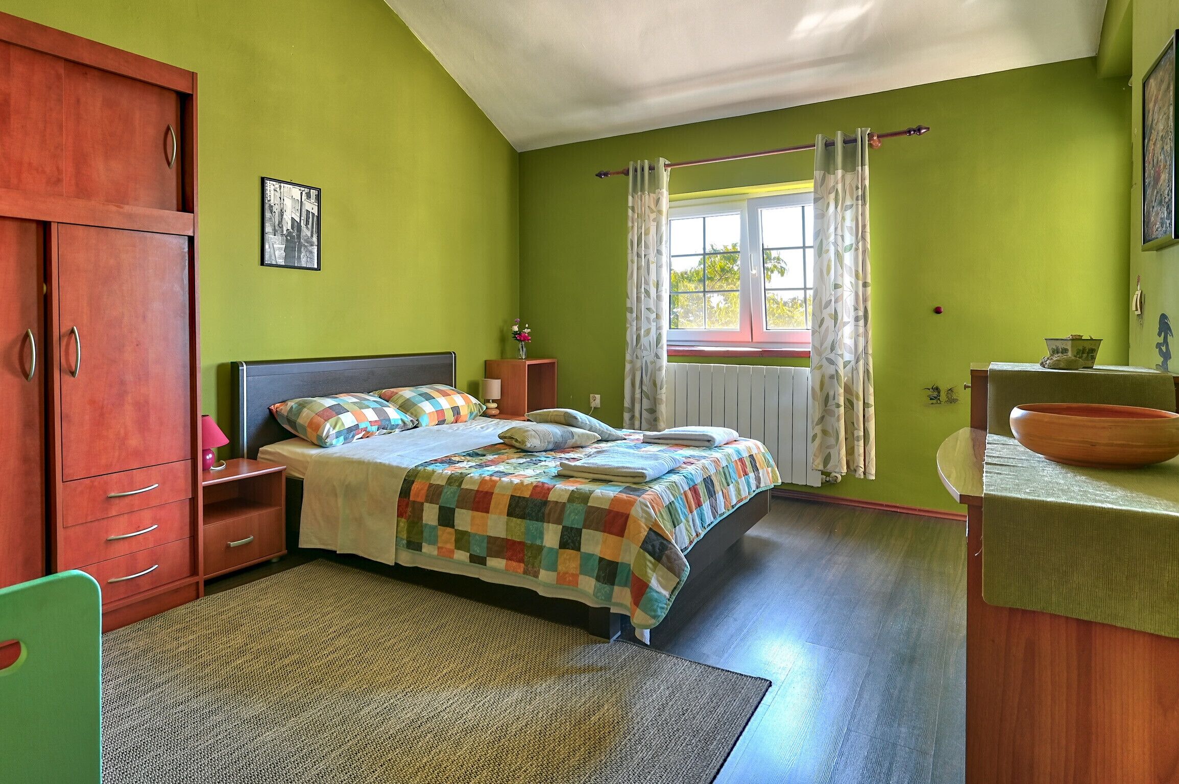 A bedroom with a bed, a wooden wardrobe, a nightstand, a window with curtains, and a bathroom with a sink and mirror.