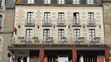 Un immeuble doté de nombreuses fenêtres et de balcons, une entrée au rez-de-chaussée avec une enseigne indiquant « LE LOU EVILLE », et des places assises à l’extérieur, protégées par des parasols.