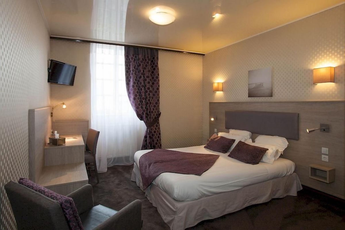 A hotel room with a bed, a desk, a chair, a TV, and a window with curtains.