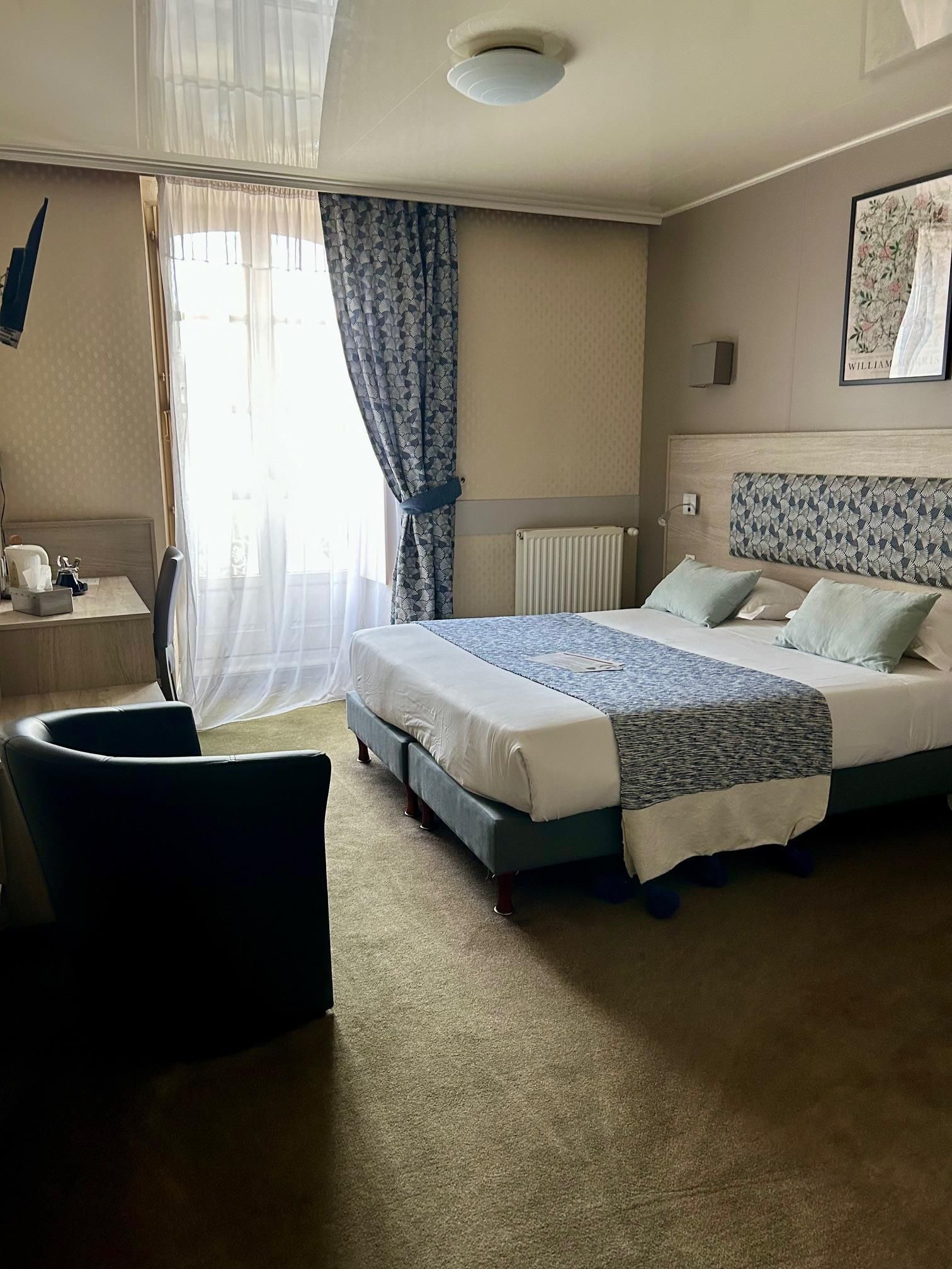 Habitación de hotel con cama, escritorio, silla, radiador y ventana con cortinas.