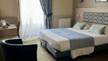 Habitación de hotel con cama, escritorio, silla, radiador y ventana con cortinas.