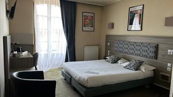A hotel room with a large bed, a desk with a chair, a radiator, and two framed pictures on the wall.