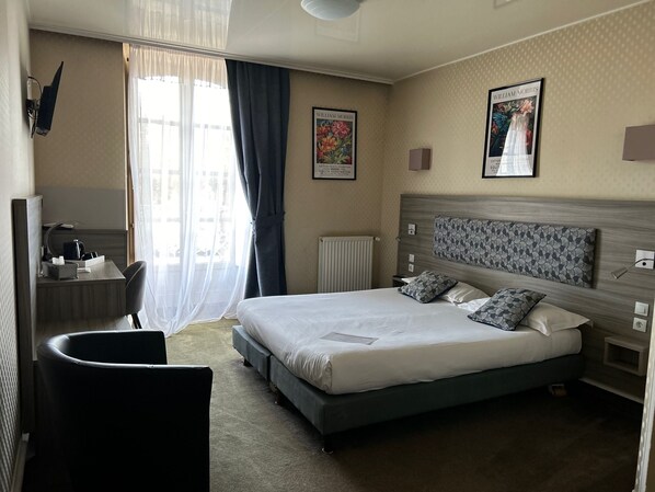 A hotel room with a large bed, a desk with a chair, a radiator, and two framed pictures on the wall. - Marguerite (Dinan)