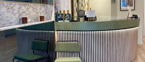 Una zona de recepció amb un taulell corbat, dues cadires verdes, una paret de marbre i un mur amb un disseny decoratiu al fons.