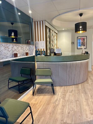 A reception area with a curved counter, two green chairs, a marble wall, and a patterned wall behind the counter. - Marguerite (Dinan)