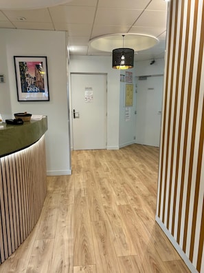 Zona de recepción con un mostrador de madera, un cuadro enmarco de un paisaje urbano, una puerta con un aviso y una lámpara colgante.