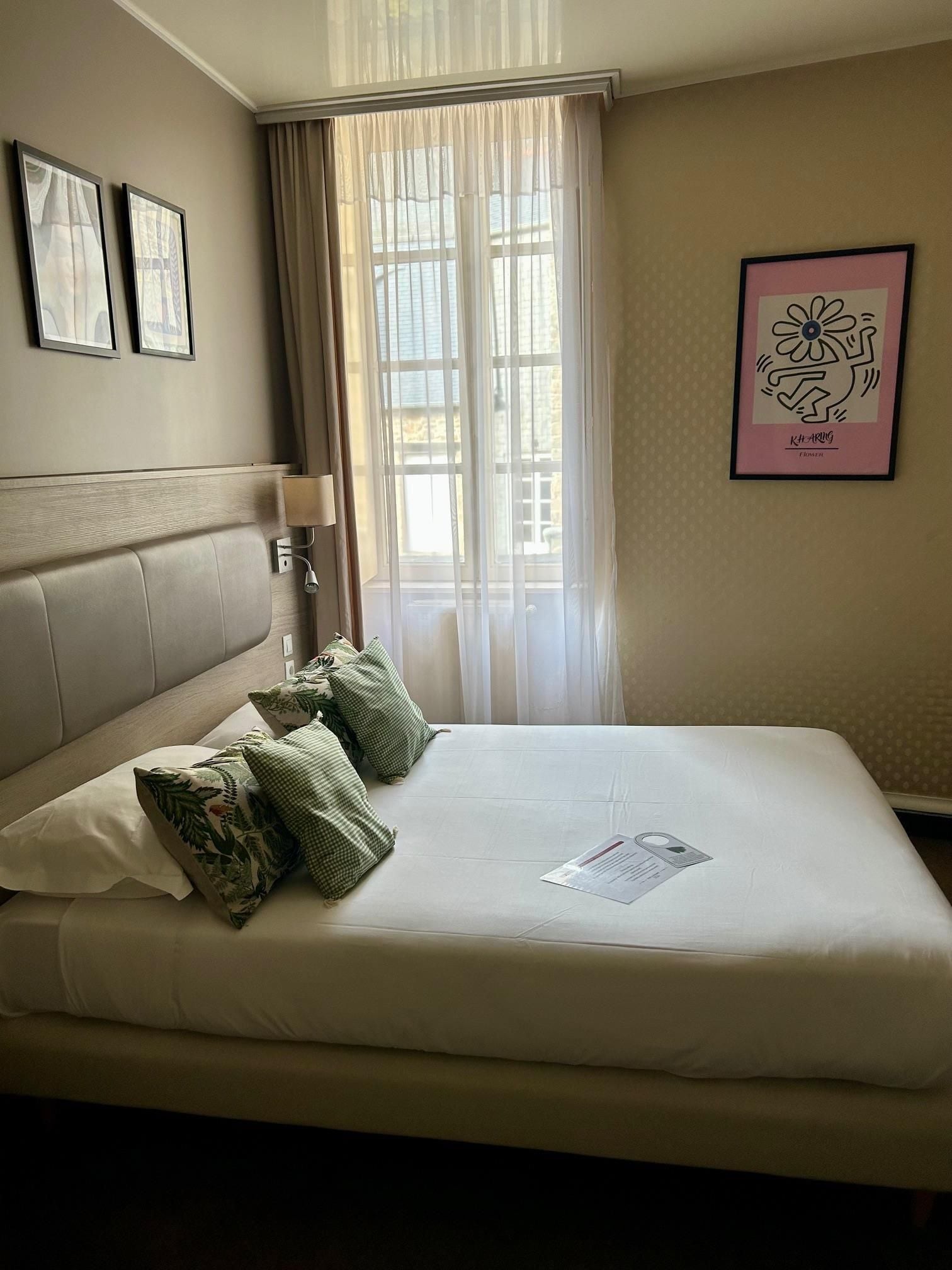 A hotel room with a bed, a headboard, a window with curtains, a wall-mounted lamp, and framed artwork.