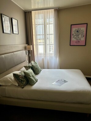 A hotel room with a bed, a headboard, a window with curtains, a wall-mounted lamp, and framed artwork. - Marguerite (Dinan)