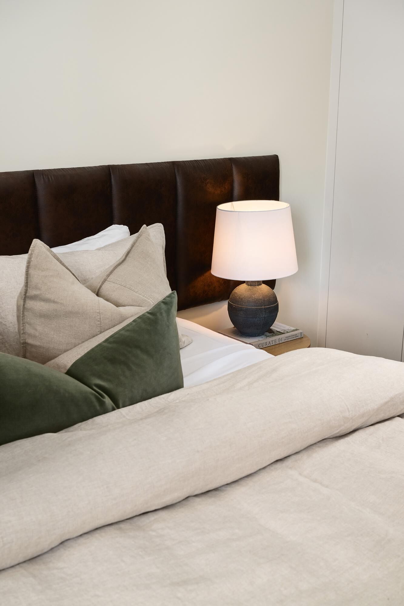 A neatly made bed with a beige comforter and pillows, a dark brown upholstered headboard, and a table lamp with a white shade.
