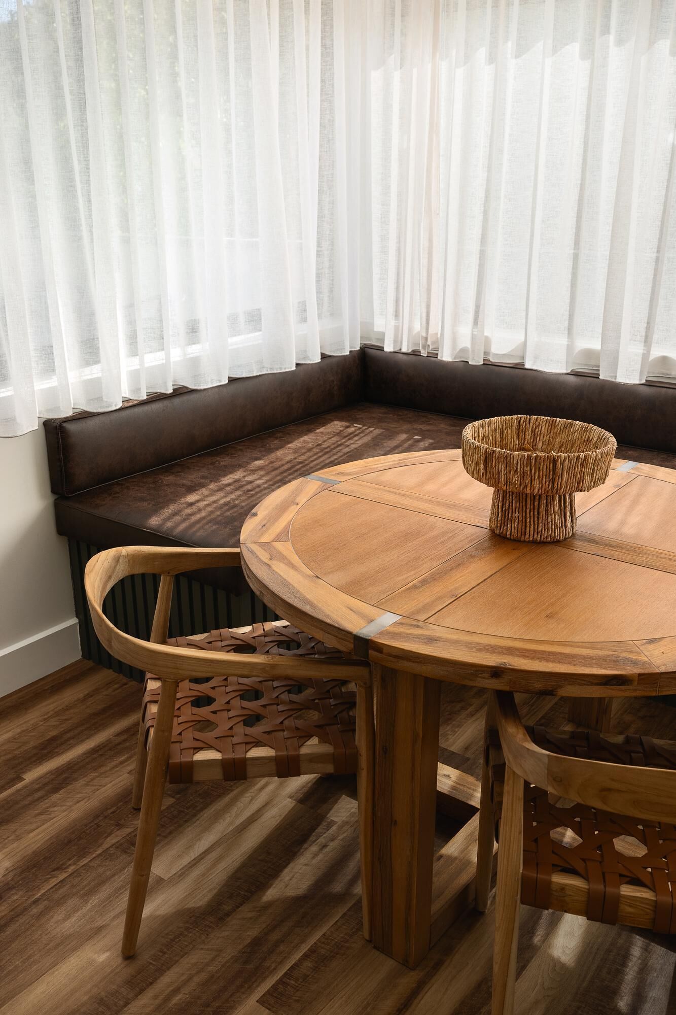 A wooden round table with a woven stool, a cushioned bench, and a chair in a room with wooden flooring and sheer curtains.