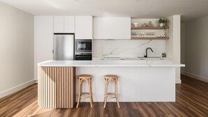A modern kitchen with white cabinetry, a built-in oven, and a marble backsplash. There are two wooden stools and a round wooden sideboard. - Apartment Two - Apartments on Gavan - Welcome to Your Luxurious Getaway at Apartments on Gavan. (Bright)