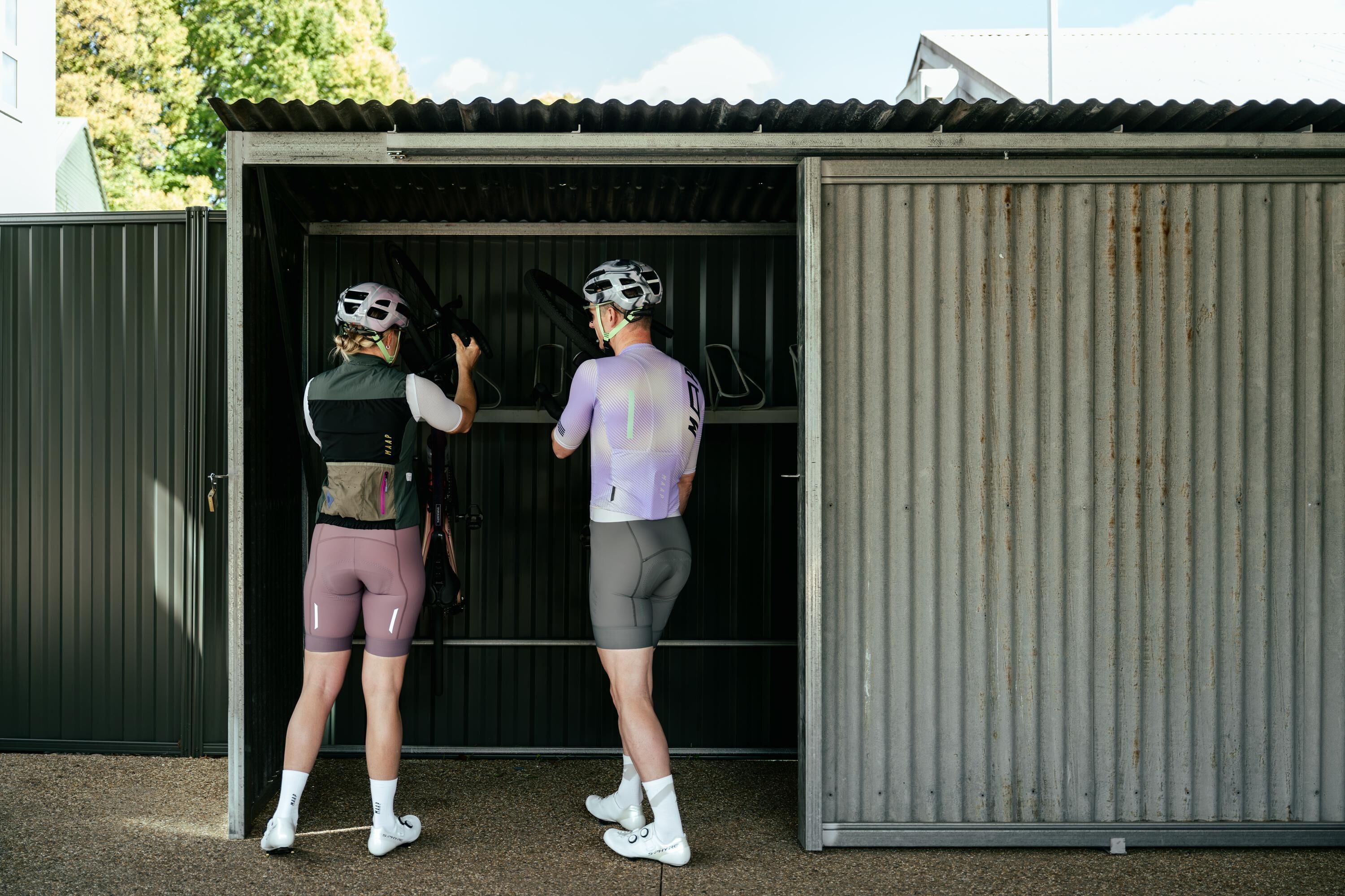 Two people in cycling gear standing in front of a metal building with a corrugated roof.