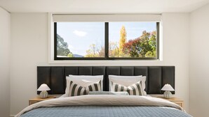 A bedroom with a large window, a bed with a quilted blanket, two bedside lamps, and a dark headboard. - Apartment Two - Apartments on Gavan - Welcome to Your Luxurious Getaway at Apartments on Gavan. (Bright)