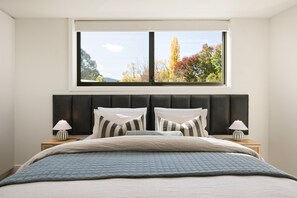 Un dormitorio con un ventanal grande, una cama con colcha acolchada, dos lámparas de noche y un cabecero oscuro.