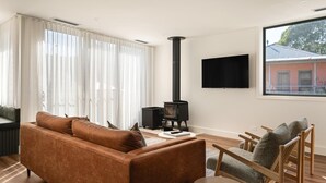 A modern living room with a sofa, chairs, a TV, and a wood-burning stove. - Apartment Two - Apartments on Gavan - Welcome to Your Luxurious Getaway at Apartments on Gavan. (Bright)