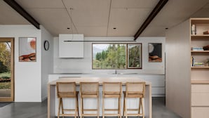A modern kitchen with a central island, wooden chairs, a built-in bookshelf, and a large window offering a view of trees. - Silver House, Ground Floor - Mt Buffalo Rd - Alpine Haven on Mount Buffalo Road – A Spacious Retreat for Families, Friends & Nature Lovers (Porepunkah)