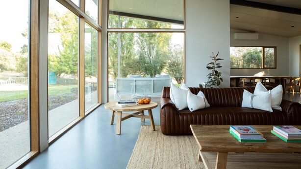 A modern living room with large windows, a brown leather sofa, a wooden coffee table, and books. - Silver House, Ground Floor - Mt Buffalo Rd - Alpine Haven on Mount Buffalo Road – A Spacious Retreat for Families, Friends & Nature Lovers (Porepunkah)