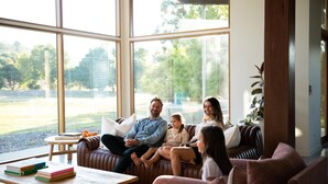 A modern living room with large windows, a leather sofa, a coffee table with books, and a view of a garden. - Silver House, Ground Floor - Mt Buffalo Rd - Alpine Haven on Mount Buffalo Road – A Spacious Retreat for Families, Friends & Nature Lovers (Porepunkah)