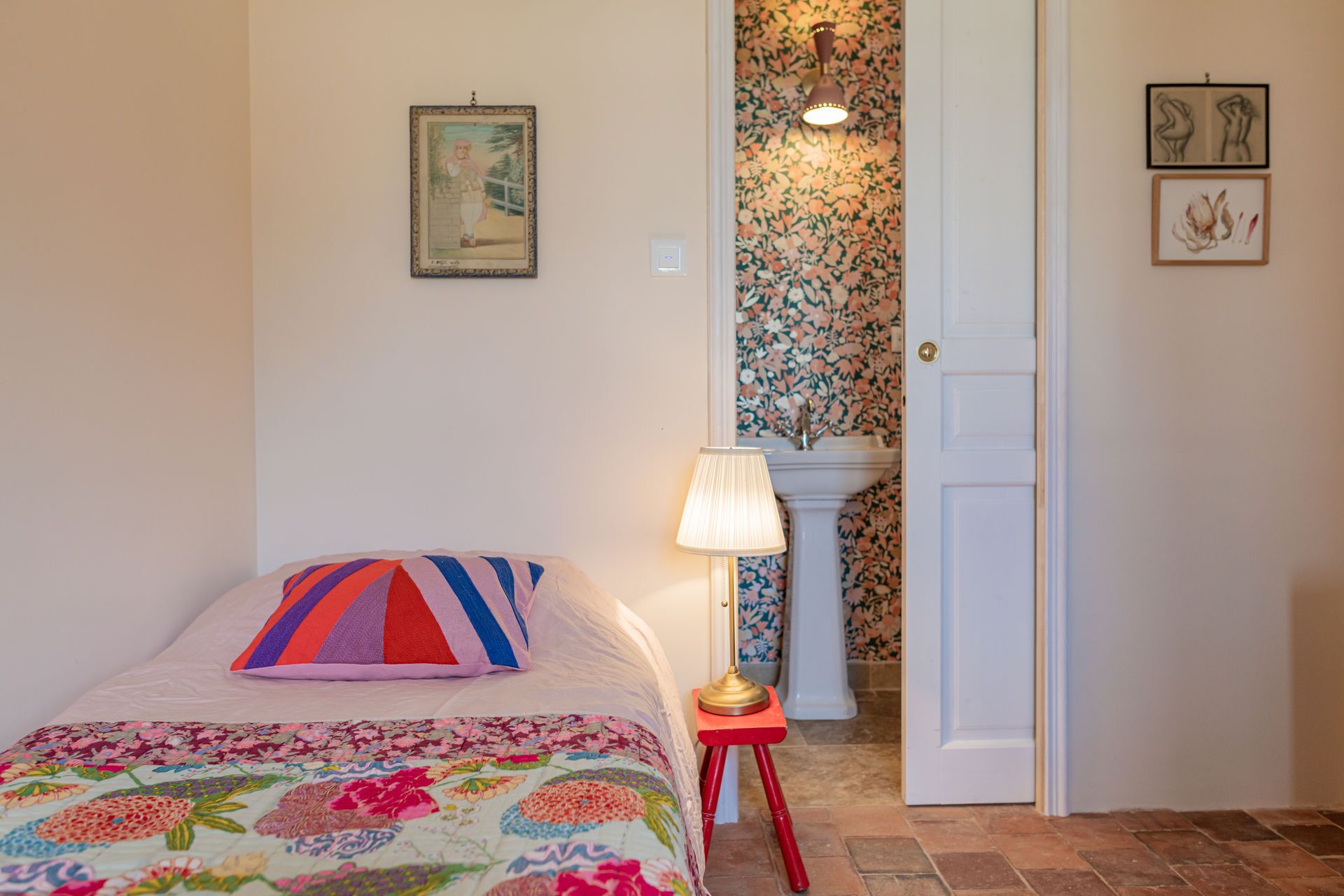 A bedroom with a floral bedspread, a colorful striped pillow, a standing lamp, and a door with floral wallpaper.