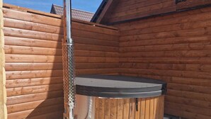 Un bain à remous en bois, doté d’une cheminée en métal, installé contre un mur en bois sous un ciel dégagé.