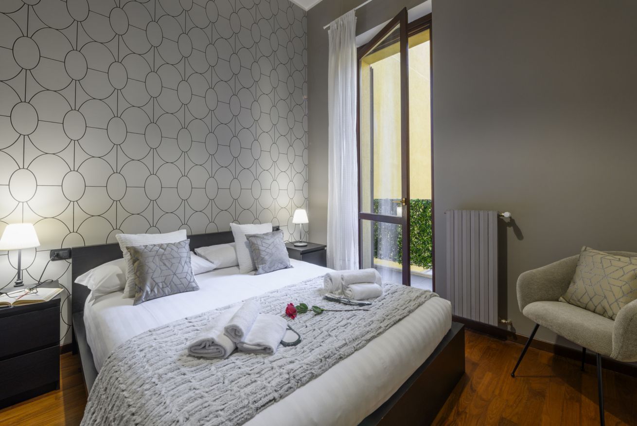 Een moderne hotelkamer met een wand met geometrisch patroon, een groot bed met witte lakens, een nachtkastje met een lamp, een stoel en uitzicht op groen door een raam.