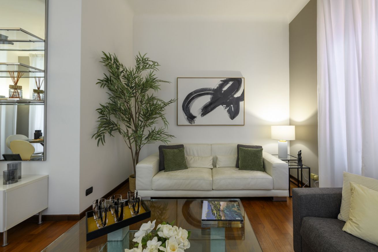 A modern living room with a white sofa, a glass coffee table, a large abstract painting, a bamboo plant, and a lamp.