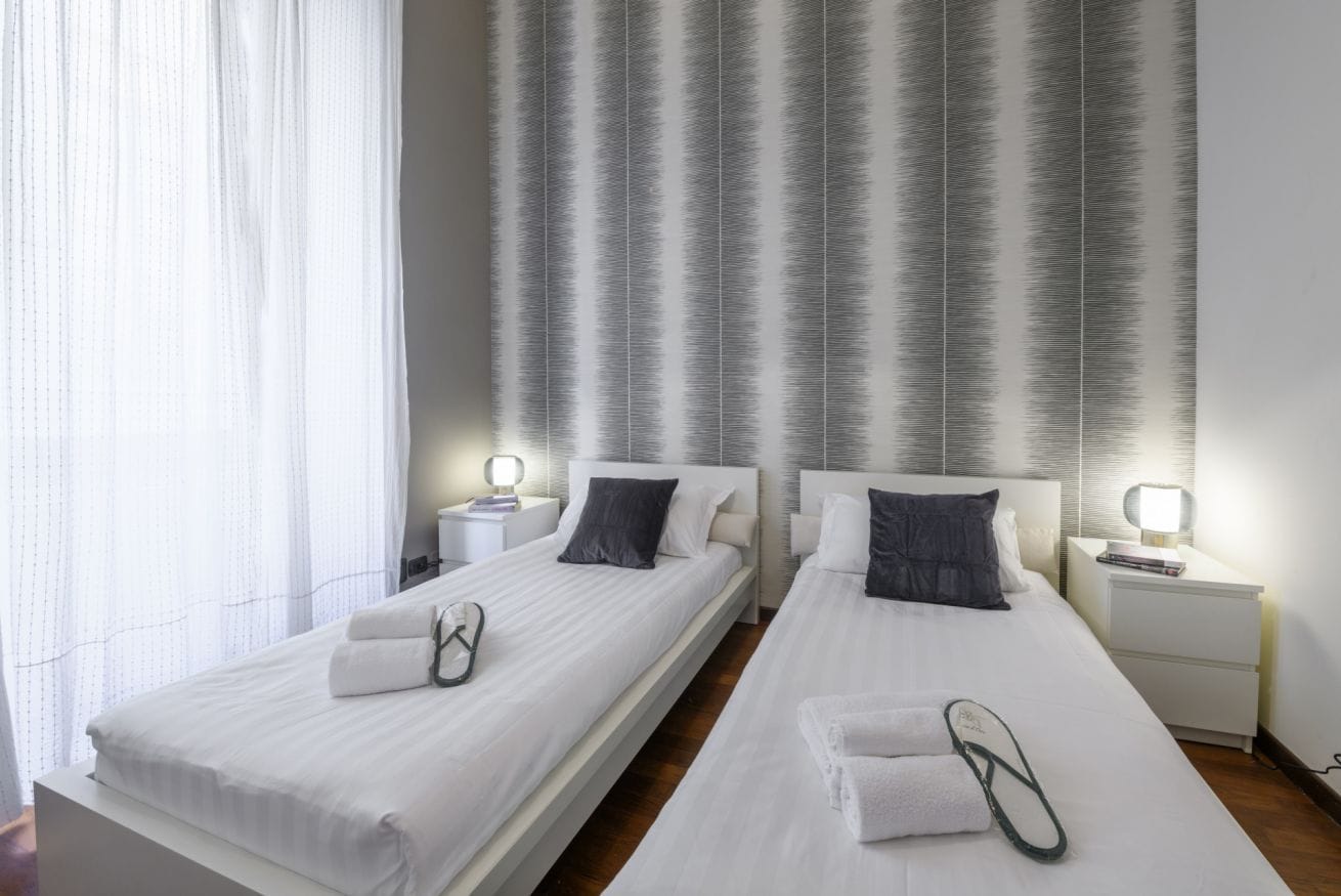 Een hotelkamer met twee bedden, witte beddengoed, donzen kussens, nachtkastjes met lampen en een raam met dunne gordijnen.