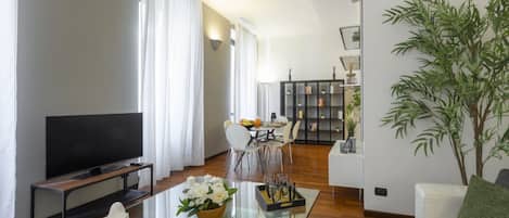 Una sala moderna con televisor de pantalla plana, una mesa de centro de vidrio con objetos decorativos, un comedor con mesa y sillas blancas, y una estantería con libros y adornos.