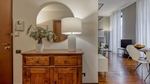 Un aparador de madera con espejo, una planta en maceta y una lámpara en una habitación con piso de madera y paredes blancas.