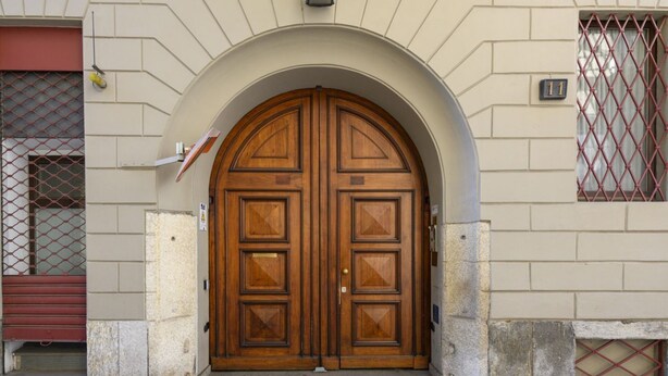 Una puerta doble de madera con un arco semicircular encima, ubicada en un edificio de paredes de ladrillo color beige y una ventana con diseño de cuadrícula roja.