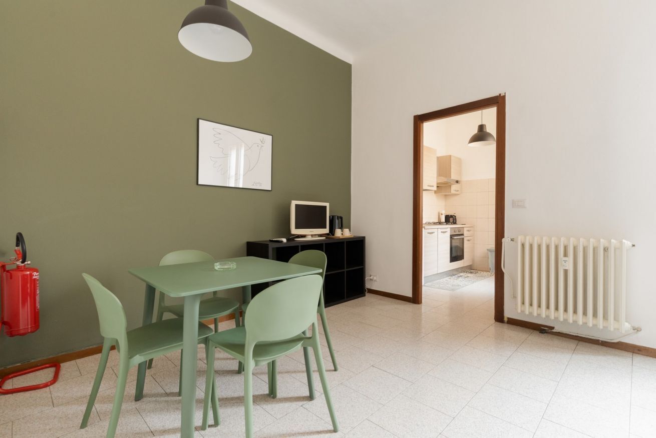 Zona pranzo con tavolo e sedie, estintore, televisore su un mobile e cucina visibile attraverso una porta aperta.