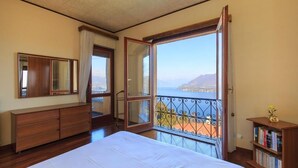 A bedroom with a large bed, a dresser, a bookshelf, and a balcony with a view of the sea.