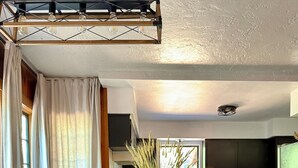 Un comedor con una mesa y sillas de madera, un jarrón con plantas secas, una cocina al fondo y una ventana con cortinas.
