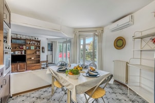 A dining area with a table set for two, a kitchen with wooden cabinets and a built-in oven, and a view of the outdoors through large windows.