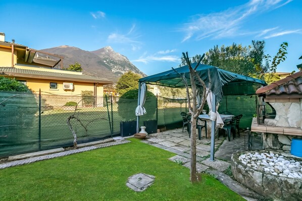 A patio with a table and chairs, a covered seating area, a stone fountain, and a mountain in the background. - Thai House Private Parking Near Lake (Toscolano Maderno)