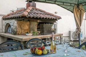 An outdoor dining area with a stone-built grill, a table set with a fruit bowl, wine glasses, and a bottle.
