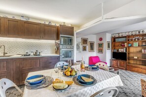 A dining area with a table set for a meal, a kitchen with wooden cabinets, and a bookshelf with trophies. - Thai House Private Parking Near Lake (Toscolano Maderno)