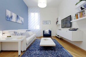 Ein modernes Wohnzimmer mit einem weißen Sofa, einem blauen Teppich mit Muster, einem weißen Couchtisch, einem blauen Sessel und einem Fernseher auf einem weißen Schrank.