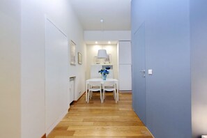 Un pasillo estrecho con piso de madera, paredes blancas y una puerta azul. Hay una mesa blanca con sillas y un jarrón de flores.