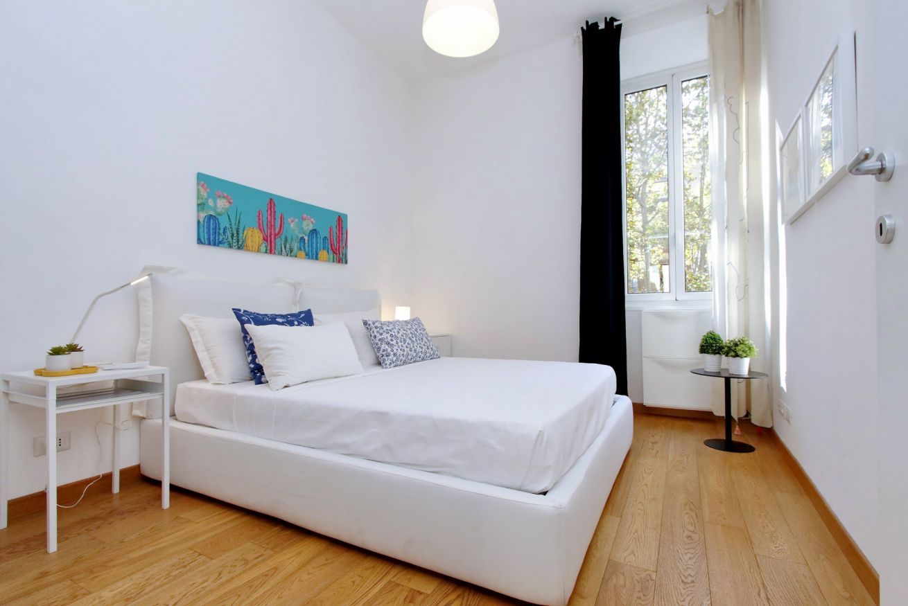 A modern bedroom with a white bed, a nightstand, a small table with a plant, and a window with curtains.