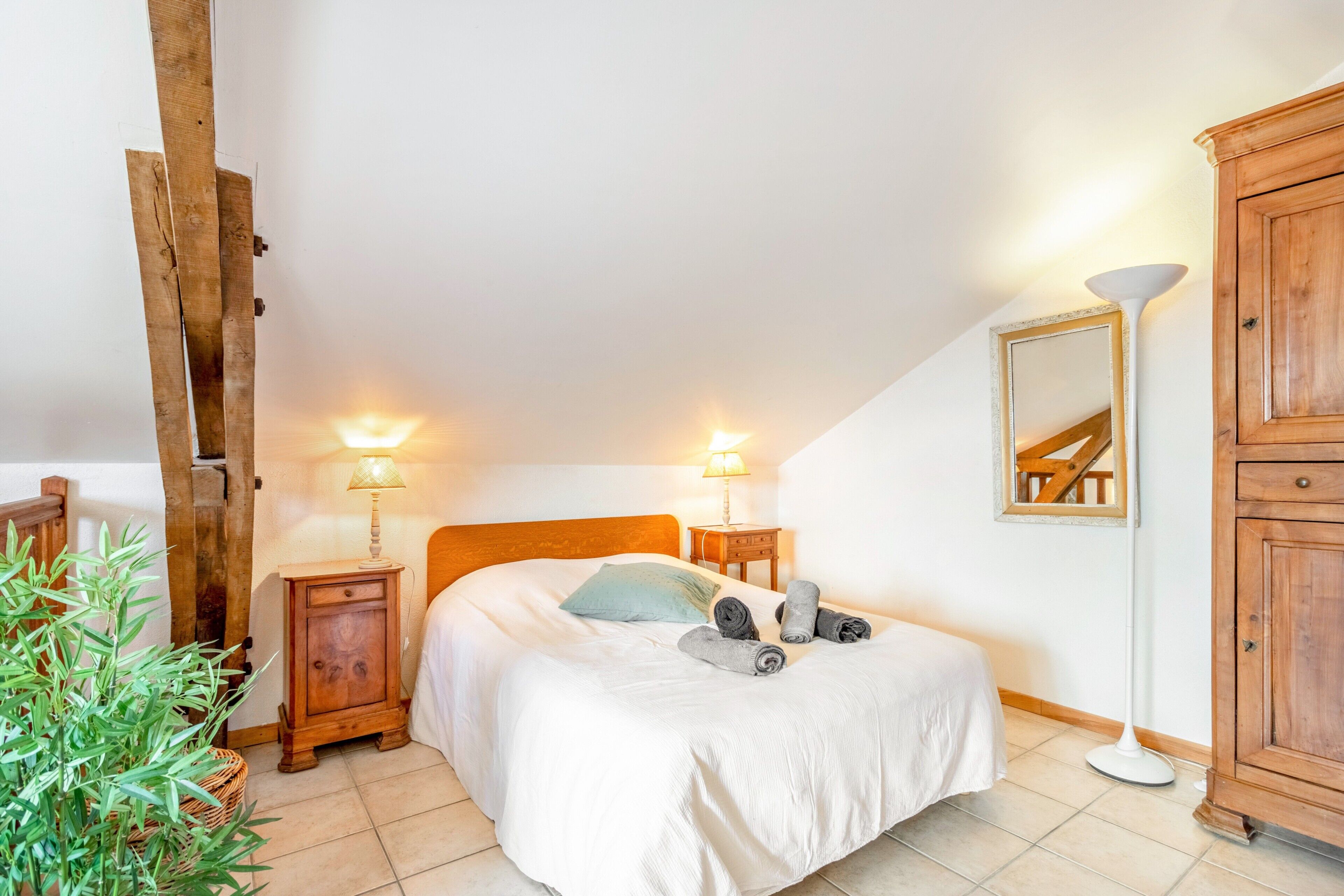Een slaapkamer met een bed, nachtlampjes, een houten garderobe, een spiegel en een potplant.