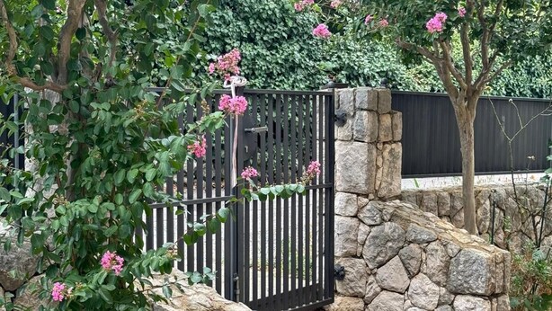 Una escalera de piedra conduce a un portón de metal negro, rodeado de abundante vegetación y flores rosadas.