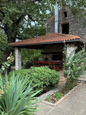 Un edificio rústico de piedra con techo de tejas, un banco de madera en el exterior y vegetación circundante.