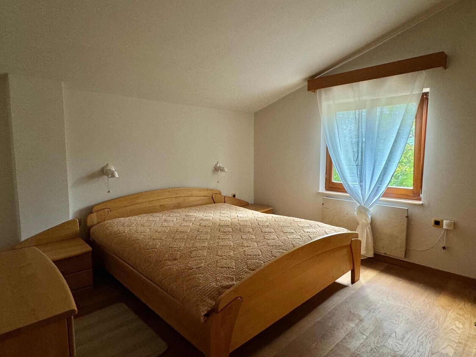 Un dormitorio con una cama de madera, una mesita de noche, una ventana con cortinas translúcidas y un radiador.